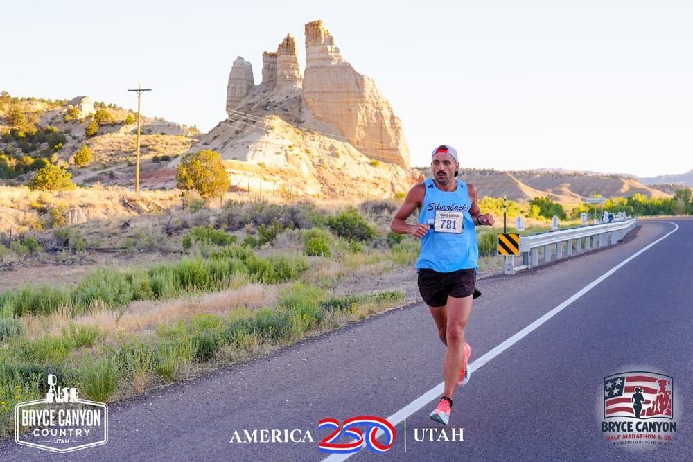 Alex T. Baker running the Bryce Canyon Half Marathon on a two-lane desert highway, light blue Silverbacks singlet, bib 78101, white cap, hoodoos and sage behind him.