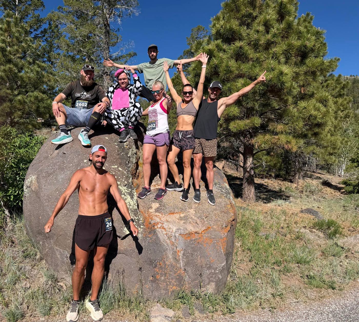 Alex T. Baker standing in front of a granite boulder in the pines, with a group of runners posed on the rock behind him after a race.